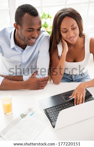 Surfing web together. Top view of beautiful young African couple looking at the laptop and smiling while sitting together at the table