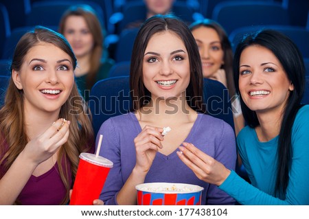 Girls at the cinema. Happy young women eating popcorn and drinking soda while watching movie at the cinema together