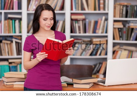 Woman in library. Cheerful young woman reading a book while leaning at the library desk