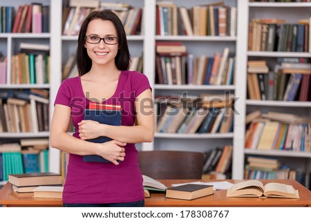 Confident and smart student. Cheerful young female student holding books and smiling while standing at the library