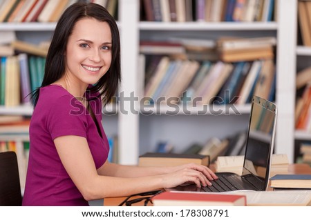 Confident student in library. Side view of cheerful young woman reading a book while sitting at the library desk