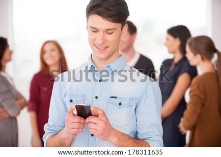 Sharing good news with friends. Cheerful young man holding a mobile phone and smiling while group of people communicating on background
