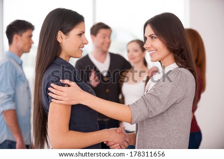 Women handshaking. Two cheerful young women handshaking while group of people communicating on background