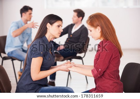 Helping each other to solve problems. Two depressed women sitting face to face and holding hands while two men communicating on background