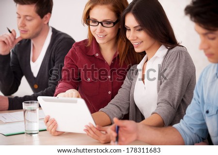 Students studying together. Group of young people sitting together at the table while two cheerful women looking at the digital tablet and smiling