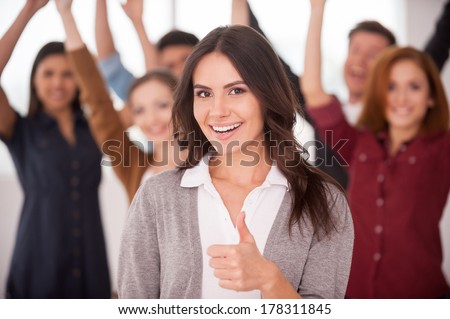 We are successful team! Beautiful young woman showing her thumb up and smiling while group of happy young people standing on background and keeping arms