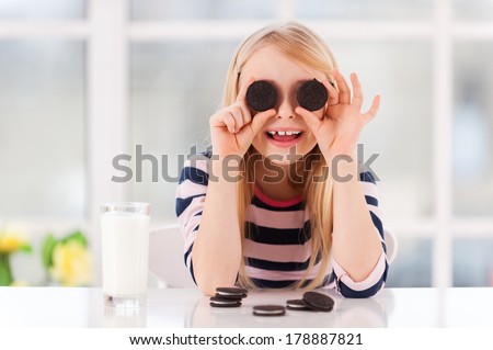 Having fun with cookies. Cheerful little girl covering eyes with cookies while sitting at the table with glass of milk on it