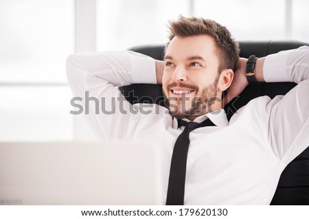 Day dreaming. Handsome young beard man in shirt and tie holding hands behind head and smiling while sitting at his working place