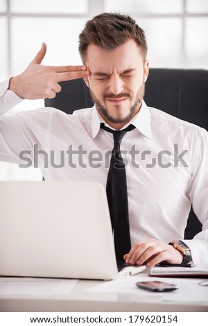Monday morning suicide. Frustrated young man in formalwear touching his temple with finger gun and keeping eyes closed while sitting at his working place