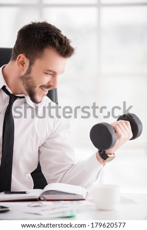 Having a sports break. Handsome young beard man in shirt and tie training with dumbbell and smiling while sitting at his working place