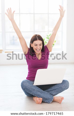 I did it! Happy young woman keeping arms raised and expressing positivity while working at laptop in her apartment