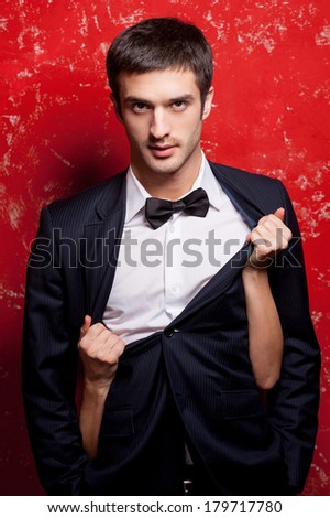 Real macho. Handsome young man in suit and bow tie standing against red background while woman hiding behind him and taking off his jacket