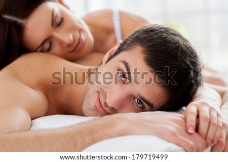 Loving couple in bed. Cheerful young loving couple lying in bed and smiling at camera