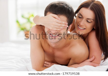 Playful loving couple. Cheerful young loving couple lying in bed while woman covering her boyfriend eyes