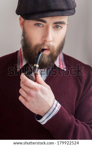 Elegance and masculinity. Portrait of handsome young man in hat smoking a pipe and smiling at camera