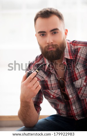 Man with a smoking pipe. Bearded handsome young man holding a smoking pipe and looking at camera while sitting on the chair
