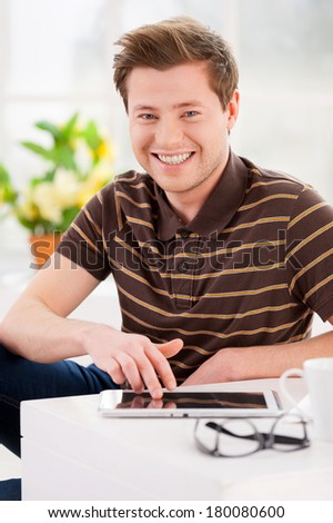 Using his new digital tablet. Cheerful young man working on digital tablet and smiling while sitting on the floor at his apartment
