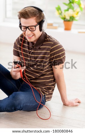 Relaxing with music. Top view of handsome young man looking at his mp3 player and listening to the music while sitting on the floor at his apartment