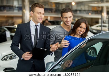 Buying their first car together. Handsome young car salesman standing at the dealership telling about the features of the car to the customers