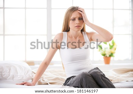 Feeling headache. Frustrated young woman touching head with hand and expressing negativity while sitting in bed at her apartment