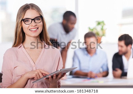 Confident in her team. Confident young woman in glasses working on digital tablet and smiling while three people working on background