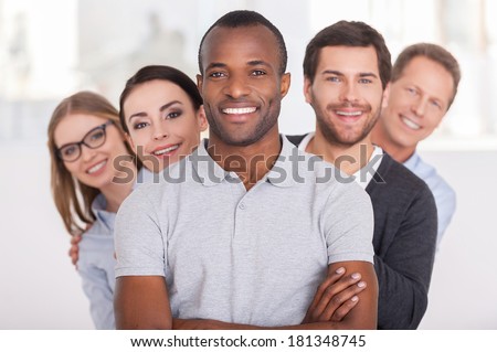 Confident business team. Cheerful young African man keeping arms crossed and smiling while group of people standing behind him in a row and looking at camera
