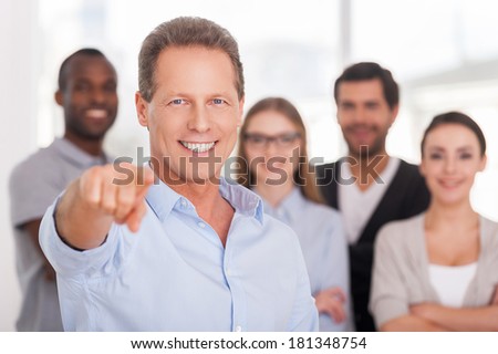 I choose you! Confident mature man pointing you and smiling while group of people in casual wear standing on background