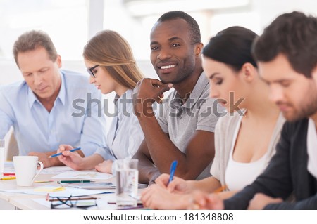 I am a part of strong and creative team. Group of business people sitting in a row at the table while handsome African man looking at camera and smiling