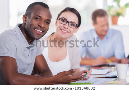 We are young and creative! Two business people sitting together at the table and smiling while other people working on background