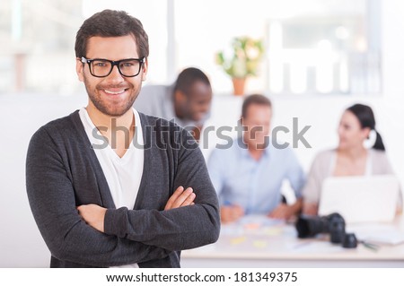 Team leader. Handsome young man in glasses keeping arms crossed and smiling while three people working on background