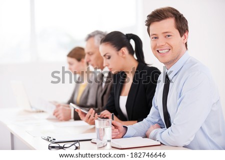 Business seminar. Side view of confident business people sitting in a row at the table while one man looking at camera and smiling