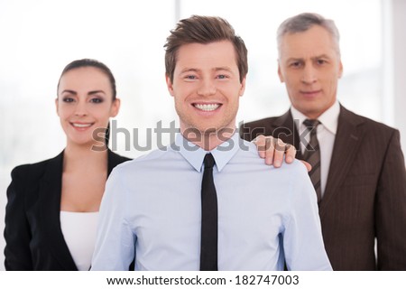 Real team leader. Cheerful young man in formalwear looking at camera and smiling while his colleagues standing behind her