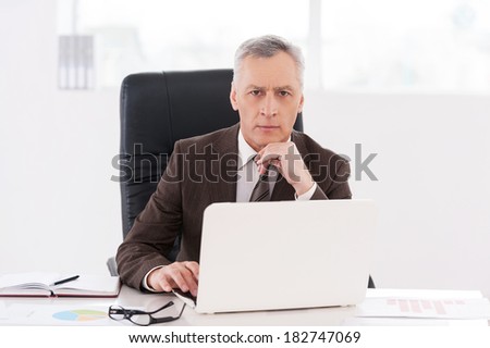 Confident businessman. Confident senior man in formalwear working on laptop and looking at camera while sitting at his working place