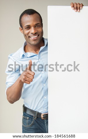 Presenting your product. Cheerful African man in blue shirt leaning at copy space and showing his thumb up while standing against grey background