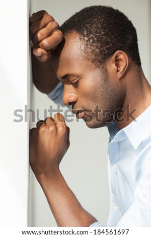 Depression and hopelessness. Side view of depressed young African man in blue shirt leaning at the wall and keeping eyes closed