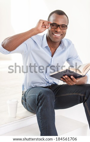 Relaxing with his favorite book. Cheerful young African man in blue shirt holding a book and looking at camera while sitting on stairs with a cup of coffee near him