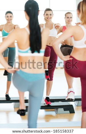 Step aerobics. Three beautiful young women in sports clothing doing step aerobics together and smiling while standing against mirror