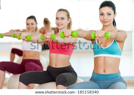Keeping their bodies in shape. Three beautiful young women in sports clothing holding dumbbells and smiling at camera while sitting on the fitness ball