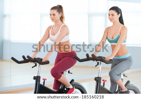 Cycling on exercise bikes. Two attractive young women in sports clothing exercising on gym bicycles