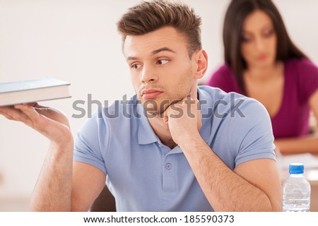 Bored student. Handsome male student holding hand on chin and looking at the textbook while young woman sitting at the desk behind him