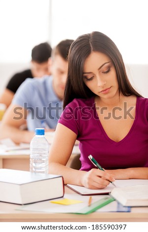 Confident student. Beautiful female student writing something in note pad while sitting in classroom with other students