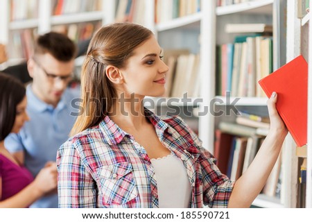 This is the book I need. Beautiful young woman picking a book from the bookshelf while people standing behind her