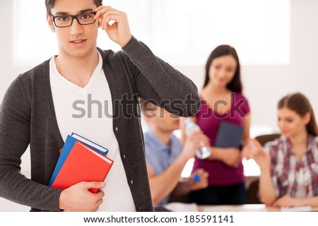 Confident and smart student. Confident male student holding books and adjusting his glasses while other students sitting on background