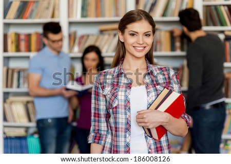 Ready for his final exam. Beautiful young woman holding books in her hand and smiling at camera while three other people standing behind her and near the bookshelf
