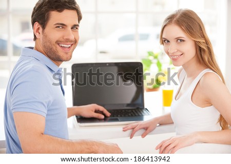 Surfing the net together. Handsome young man sitting at the table and using laptop while his girlfriend standing behind him