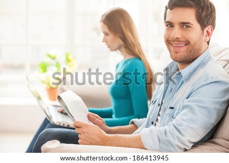Enjoying their day off together. Handsome young man sitting on the couch and holding a newspaper while his wife sitting on the background and working on laptop