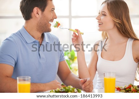 Try my meal! Beautiful young couple sitting together at the table while woman feeding her boyfriend with salad