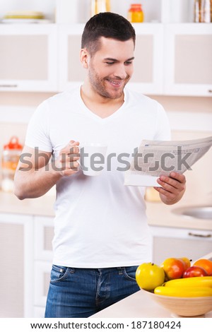Carefree morning. Handsome young man reading a newspaper and holding a coffee cup while standing in the kitchen