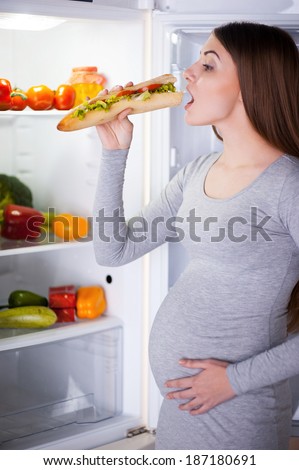 Hungry pregnant woman. Beautiful young pregnant woman standing near the open fridge and eating sandwich