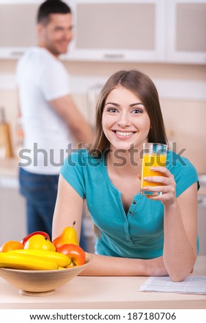 Spending Sunday morning together. Beautiful young woman leaning at the kitchen stove and holding a glass with orange juice while man standing on background and smiling